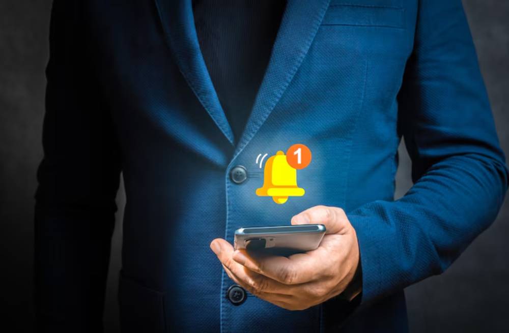 A man in a blue suit holds a smartphone displaying a notification alert with a bell icon and a '1' badge.