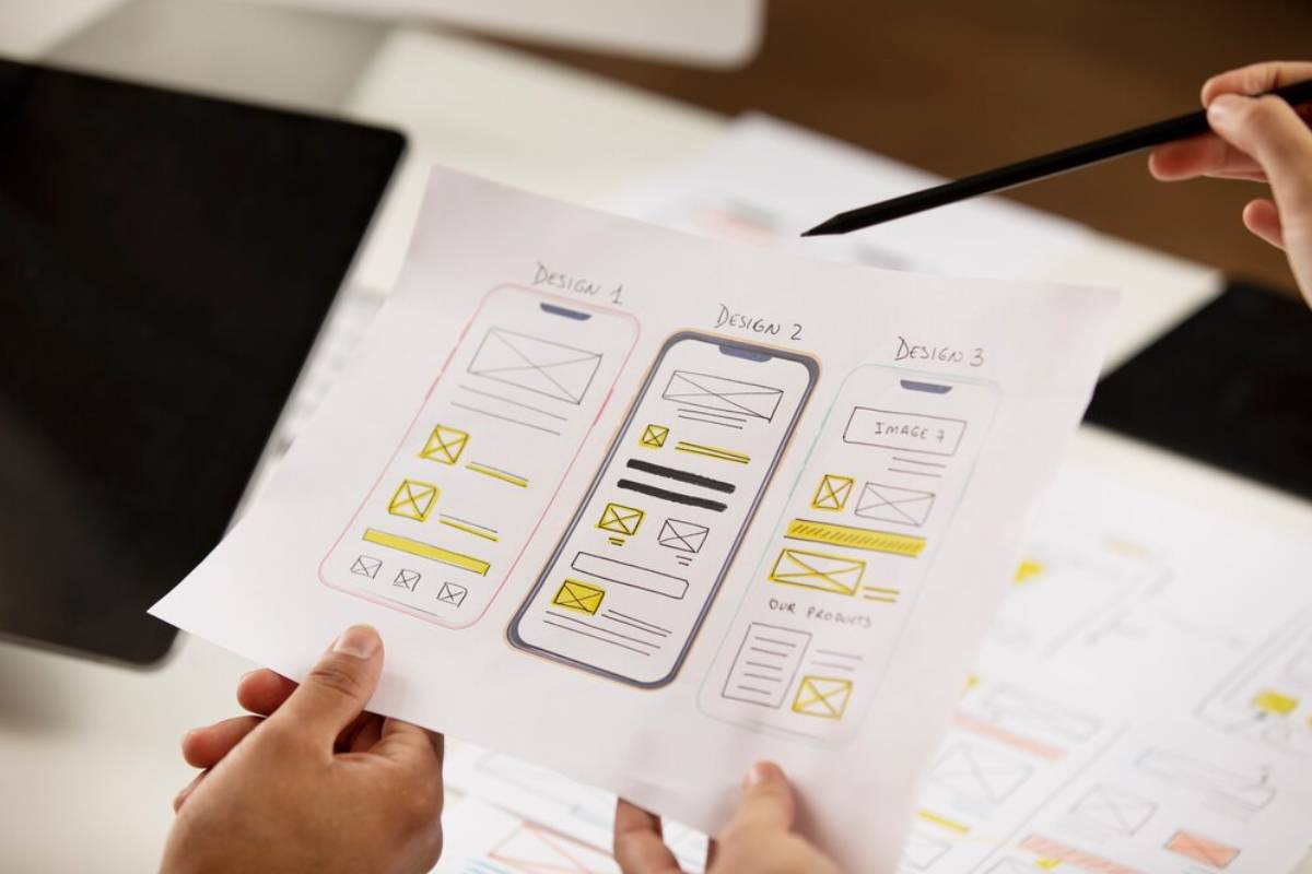 A person holding a paper showing three different mobile app design mockups, labeled Design 1, Design 2, and Design 3, with sketches.