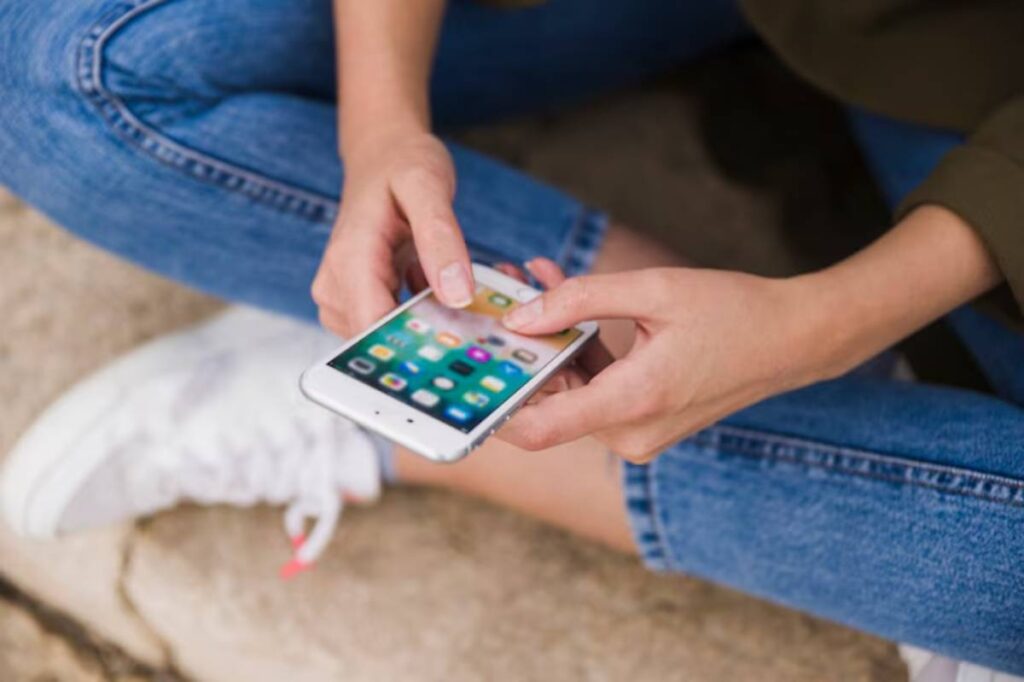 A close-up of a person’s hands using a smartphone while seated on a stone surface, wearing blue jeans and white sneakers.