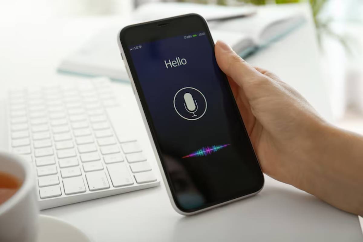 A person holds a smartphone displaying a voice recognition interface with the word 'Hello' while a keyboard and tea are nearby.