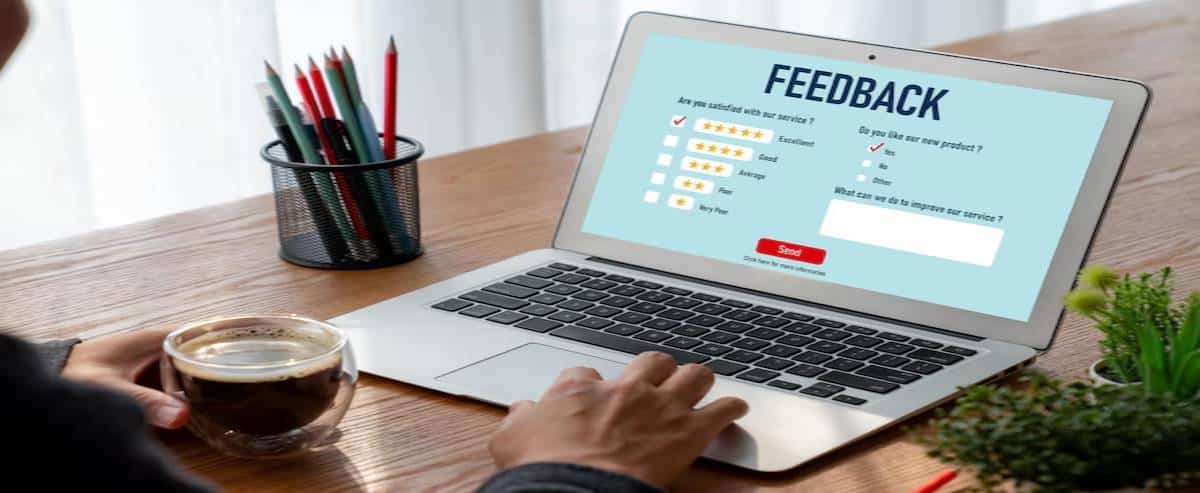 A person completes an online feedback form on a laptop, with a coffee cup and a pen holder nearby on a wooden table.