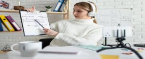 A person in a cozy sweater uses a pointer to explain future tenses on a whiteboard during an online learning session.