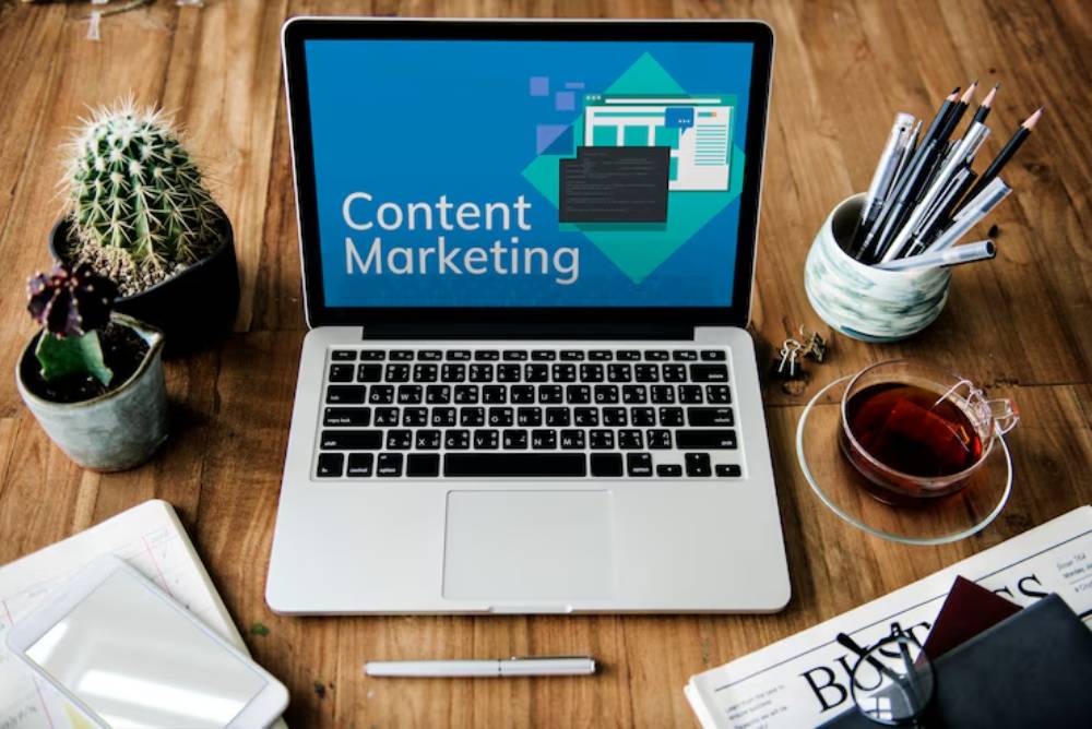 A laptop displays 'Content Marketing' on the screen, surrounded by plants, a teacup, stationery, and a smartphone on a wooden desk.