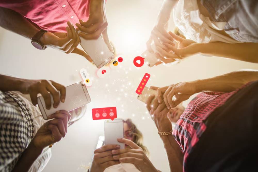 A group of people holding smartphones, with social media icons and emojis floating above them, capturing a moment of online interaction.