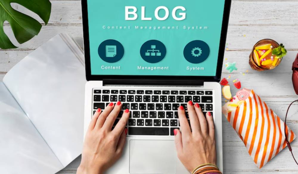 A person types on a laptop displaying a 'Blog: Content Management System' interface, surrounded by colorful snacks and a plant.