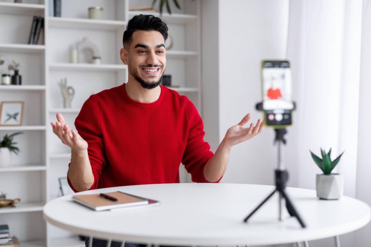 A person in a red sweater gestures while recording on a smartphone on a tripod, seated at a round table in a bright room.
