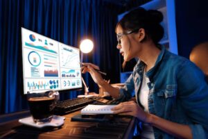 A person in a denim jacket analyzes data on a computer screen, taking notes with a pen beside a coffee cup on a desk.