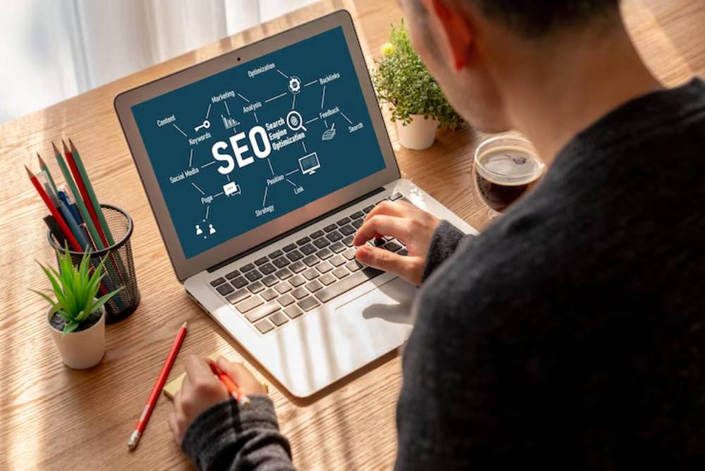 A person types on a laptop featuring an SEO mind map, surrounded by plants, a cup of coffee, and stationary on a wooden desk.