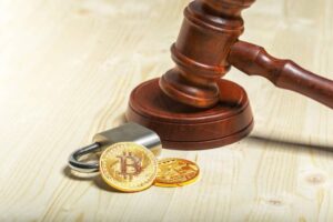 A wooden gavel rests beside a padlock and two gold Bitcoin coins on a light wooden surface, symbolizing cryptocurrency and law.
