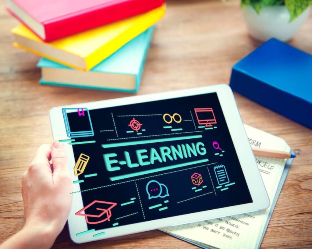 A hand holds a tablet displaying 'E-Learning' with educational icons, surrounded by colorful books and a notepad on a wooden table.