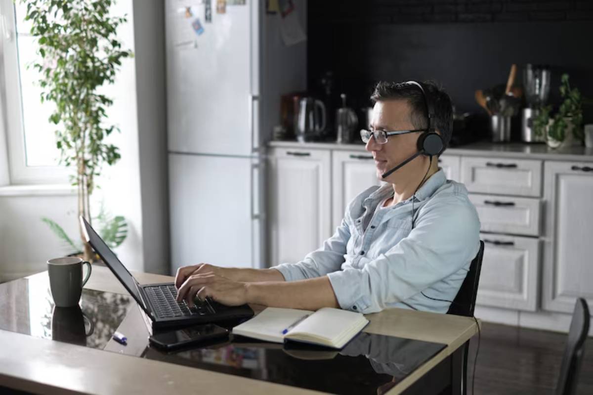 A person wearing headphones sits at a desk, typing on a laptop with a notebook and a coffee cup beside them, in a well-lit kitchen.