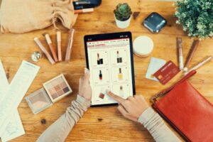 A person browses an online shopping site for cosmetics on a tablet, surrounded by makeup products, a credit card, and a purse.