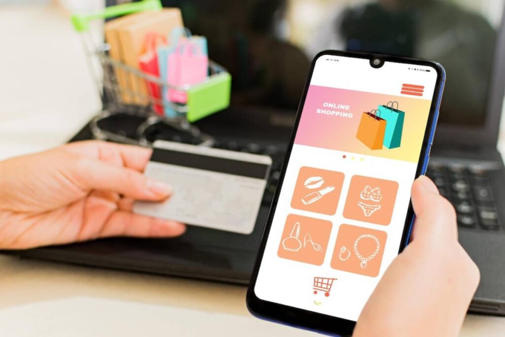 A person holding a credit card and smartphone displaying an online shopping app, with a laptop and shopping cart in the background.