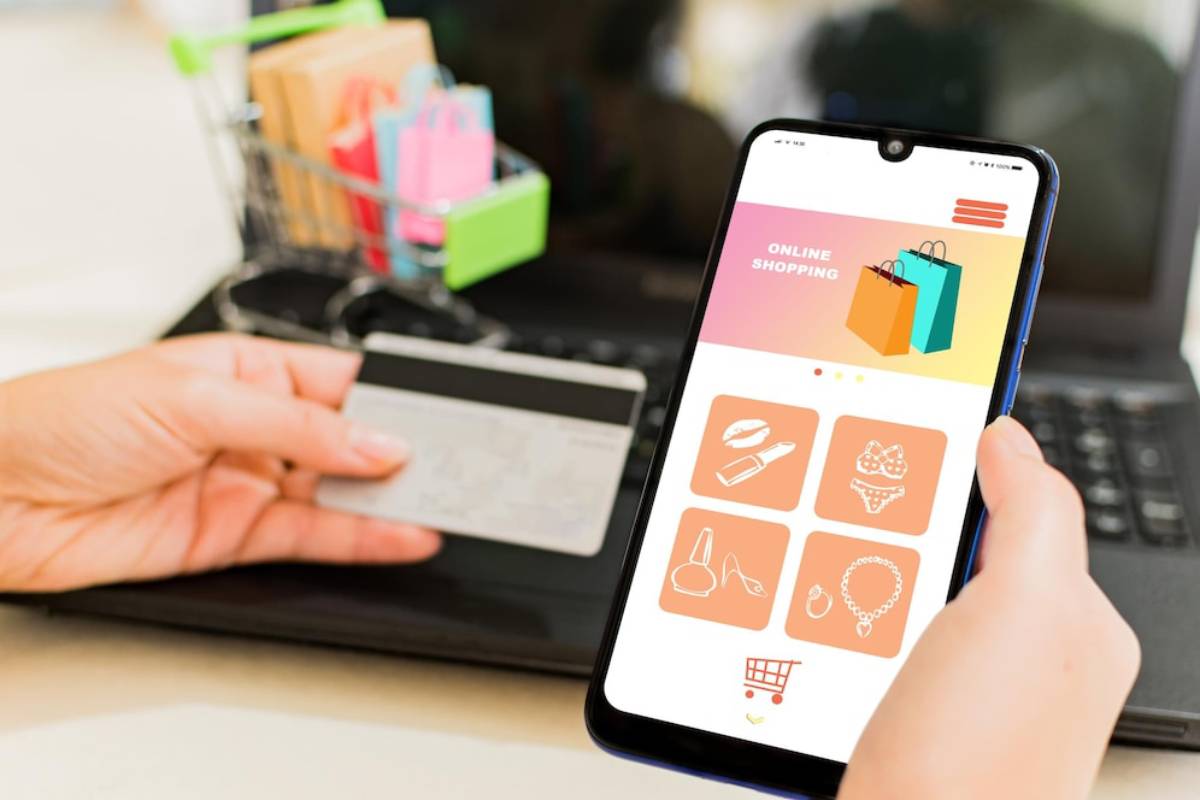 A person holding a credit card and smartphone displaying an online shopping app, with a laptop and shopping cart in the background.