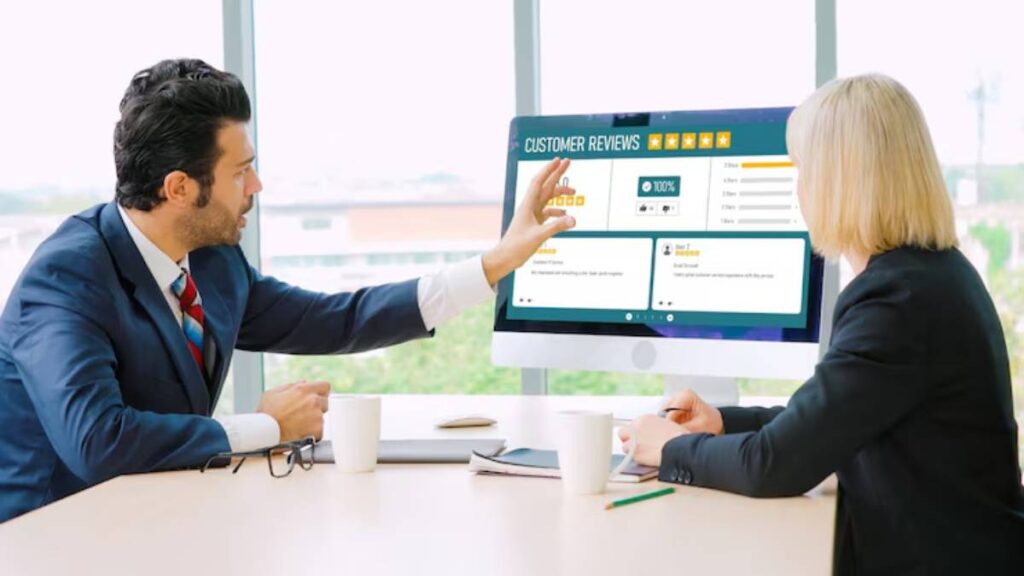 A man and a woman in business attire discuss customer reviews displayed on a computer screen, with coffee cups on the table.
