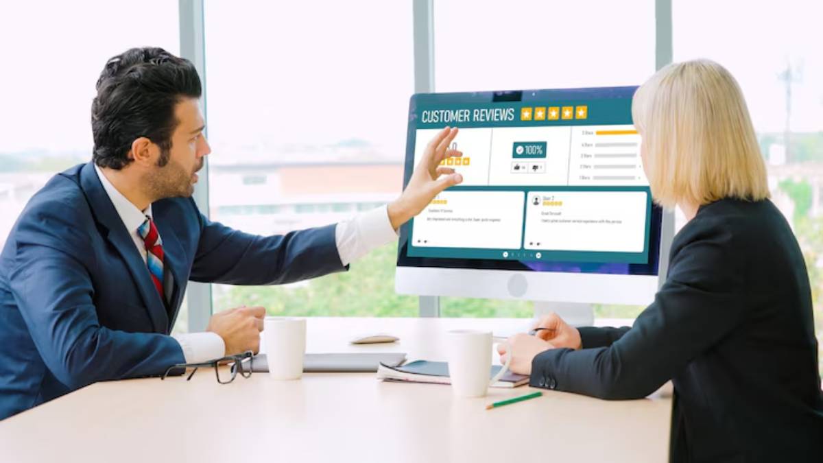 A man and a woman in business attire discuss customer reviews displayed on a computer screen, with coffee cups on the table.