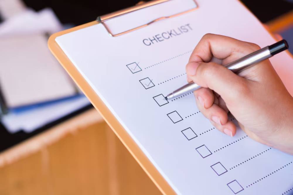 A hand holding a pen checks off items on a checklist on a clipboard, with blurred papers in the background.