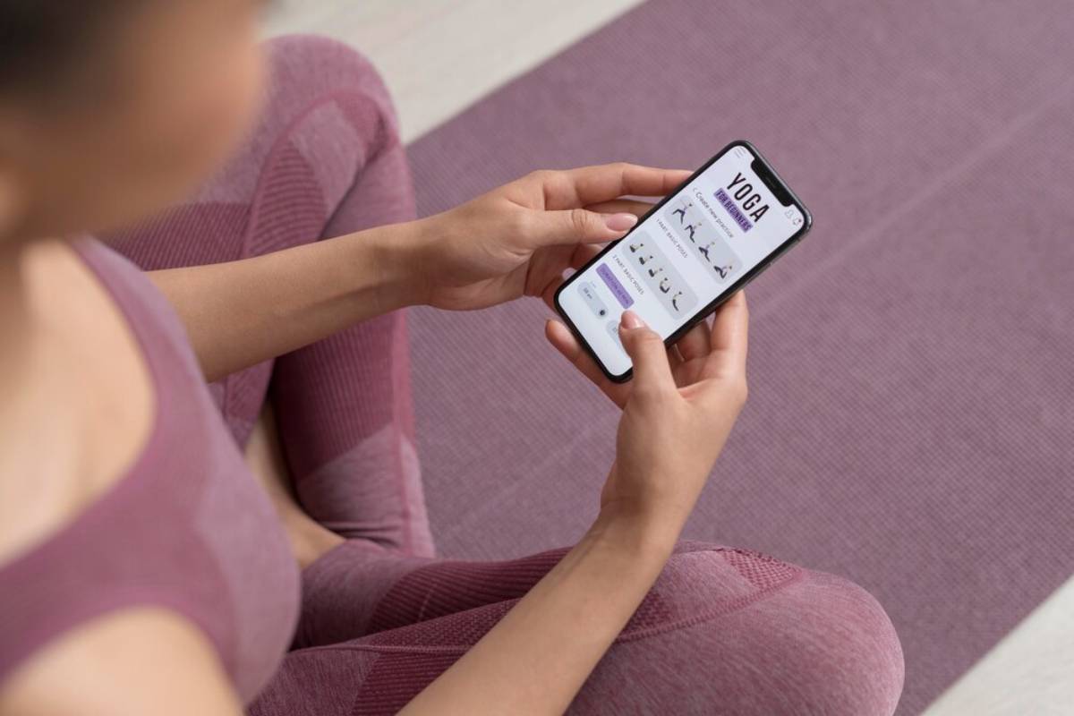 A person in a pink yoga outfit sits on a mat, holding a phone displaying a yoga app with various poses.