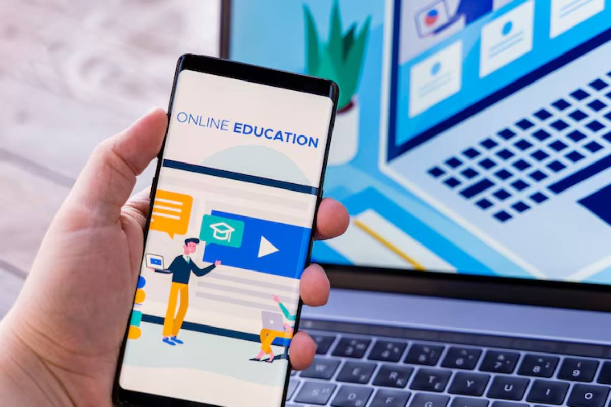 A hand holding a smartphone displaying 'Online Education,' with a laptop in the background featuring educational graphics.