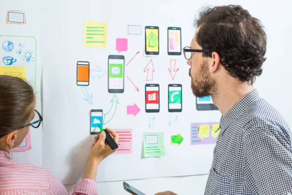 Two people collaborate on a project board filled with sketches of mobile app interfaces and colorful sticky notes.