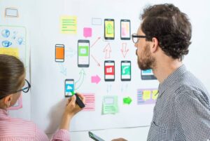 Two people collaborate on a project board filled with sketches of mobile app interfaces and colorful sticky notes.